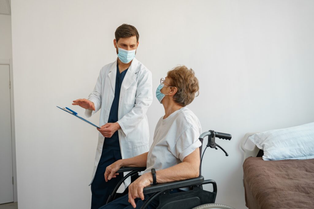 Doctor in mask explaining to a sick patient in a wheelchair the details of treatment